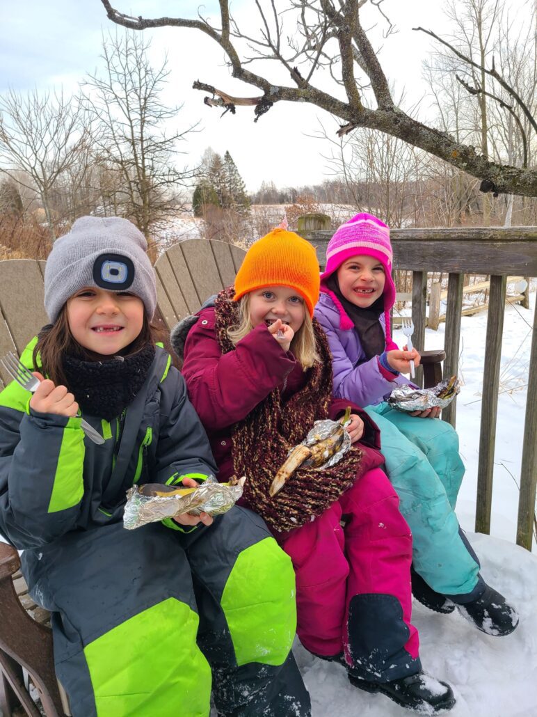 3 young campers dressed in winter gear sit on a bench outside with snow on the ground. They are smiling and holding food cooked on a campfire.