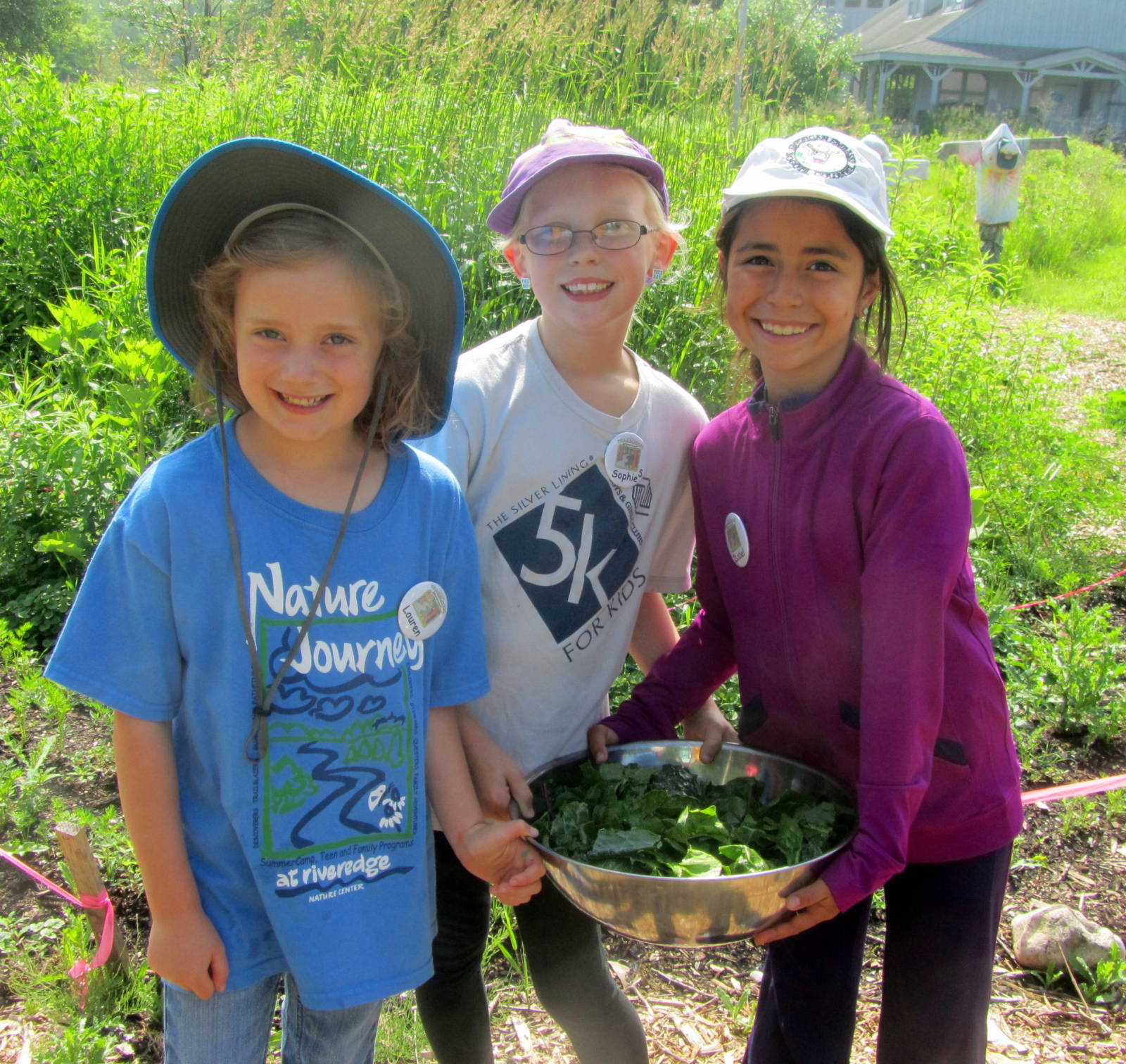 3 young kids smile at the camera while holding a bowl of veggies in the Riveredge Children's Garden