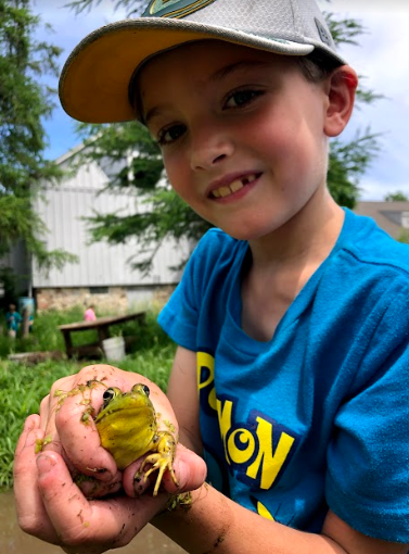 a young boy smiles at the camera holding a frog outside in the Riveredge yard