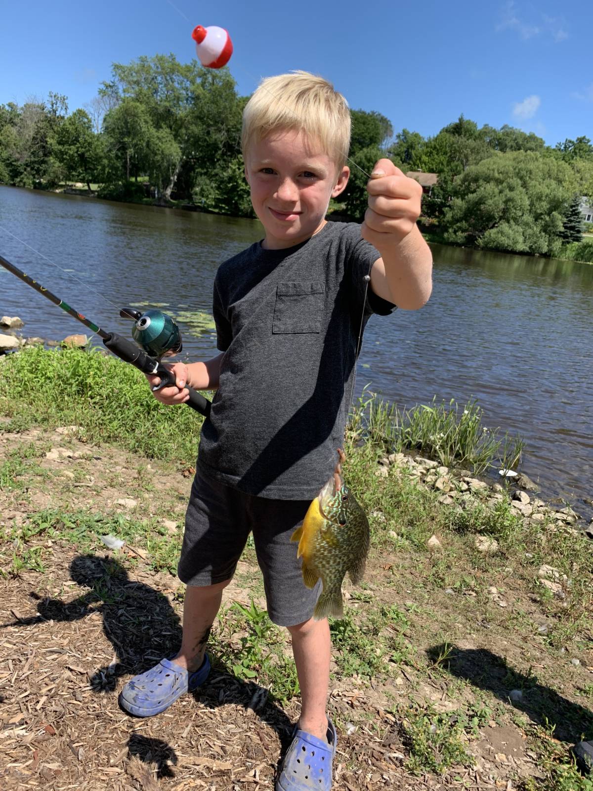a young boy proudly holds up a small fish at the end of his line. The river is behind him.