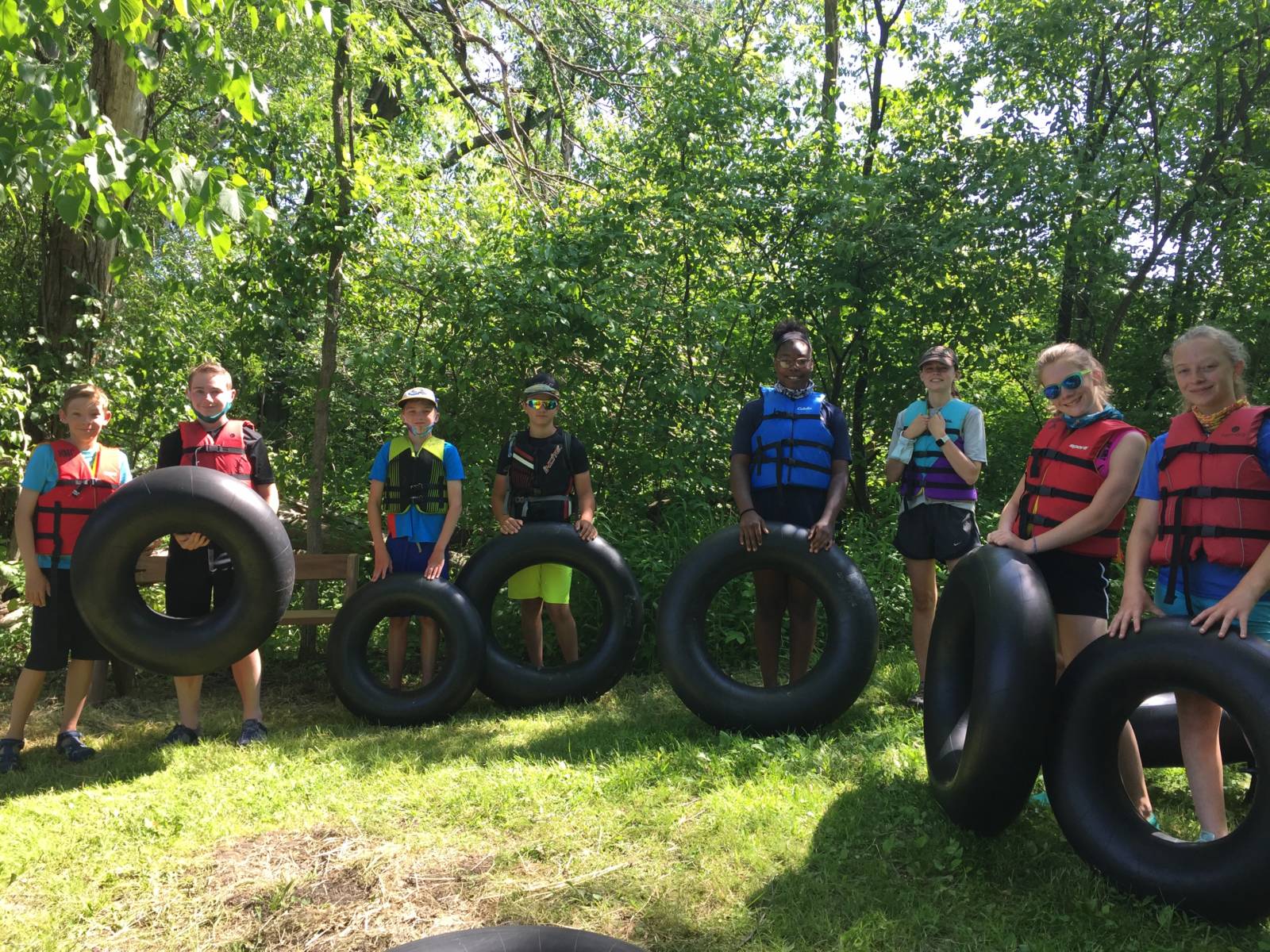 A group of kids wearing PFDs and holding inner tubes with trees behind them