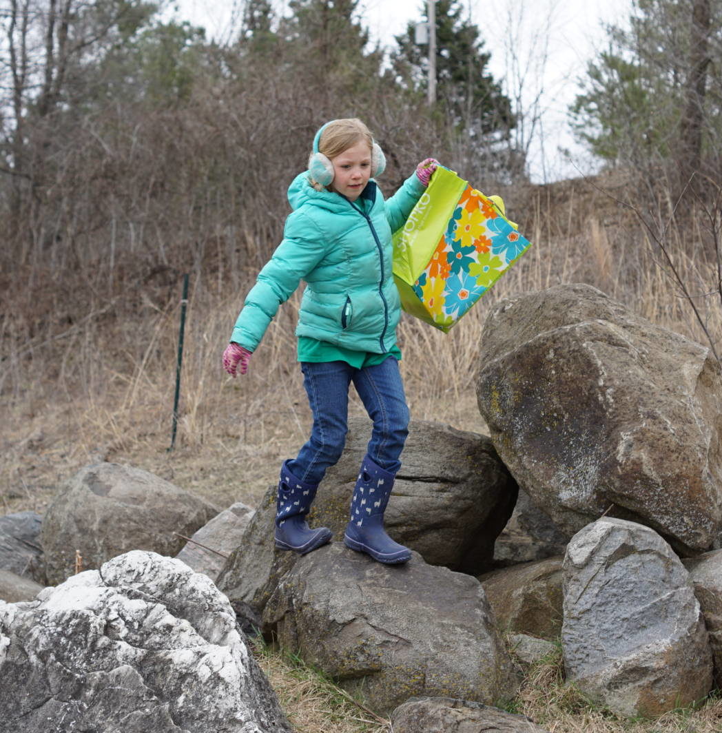 a child stands on the Larsen Climbing Rocks at Riveredge on a cold spring day holding a bag to collect eggs at the spring egg hunt