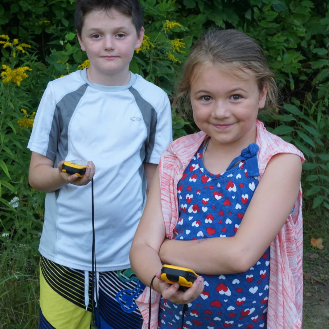 2 kids standing on a wooded trail using GPS devices during a geocaching class