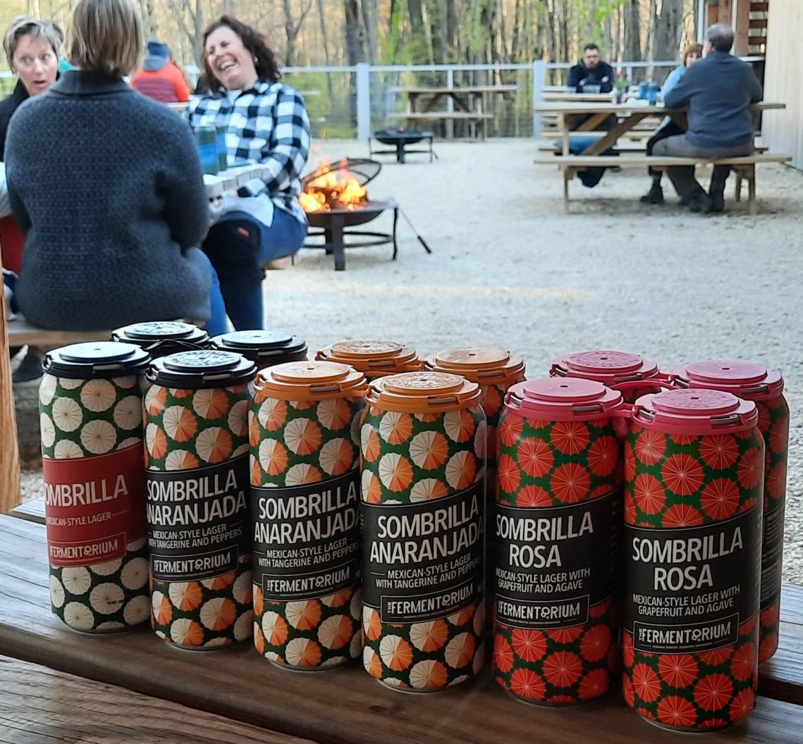beers from the fermentorium lined up on a table with people sitting outside at the Riveredge Sugarbush House in the background