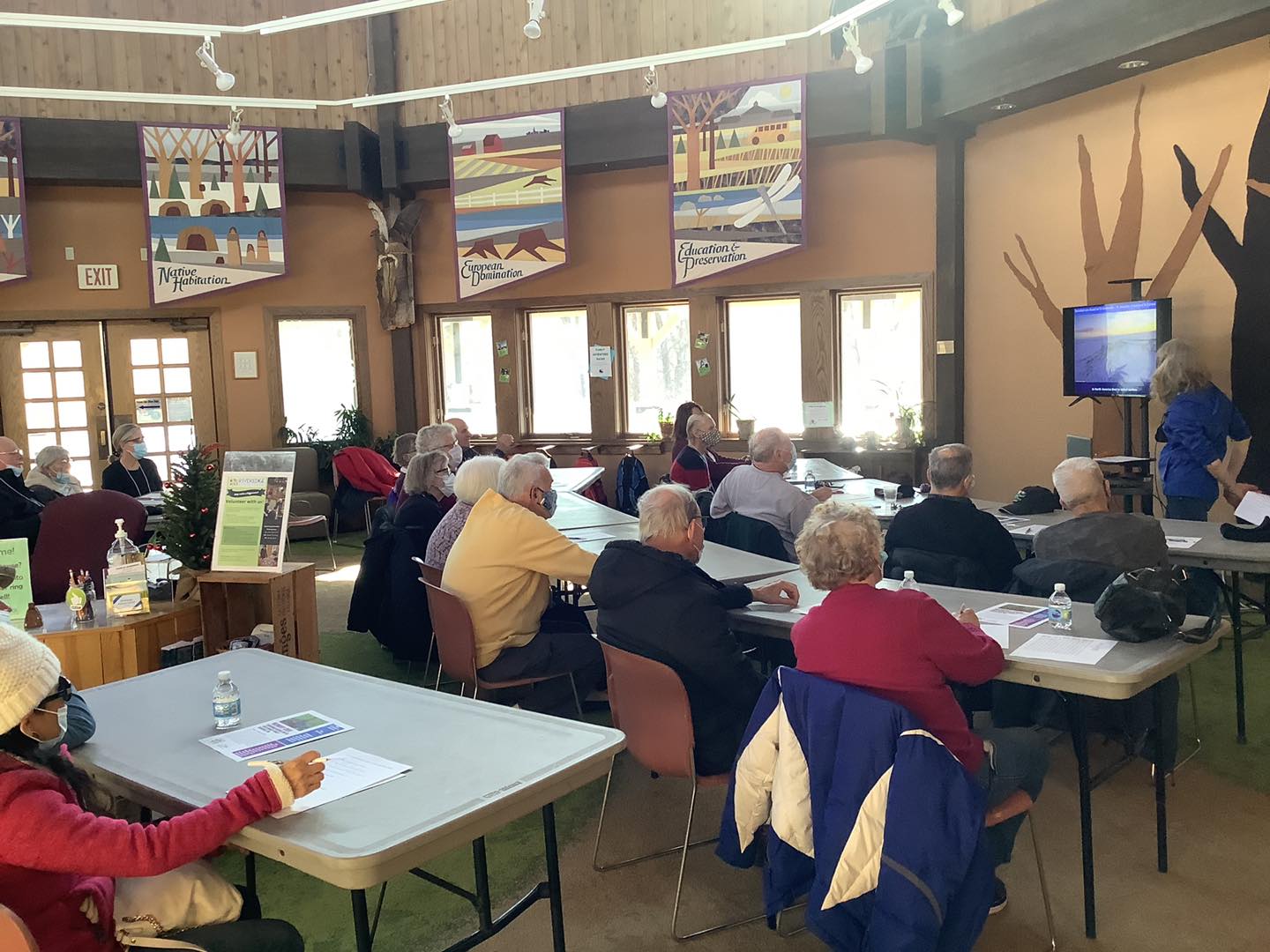 a group of memory cafe adults sit in the Riveredge visitor center watching a presentaiton by Mary