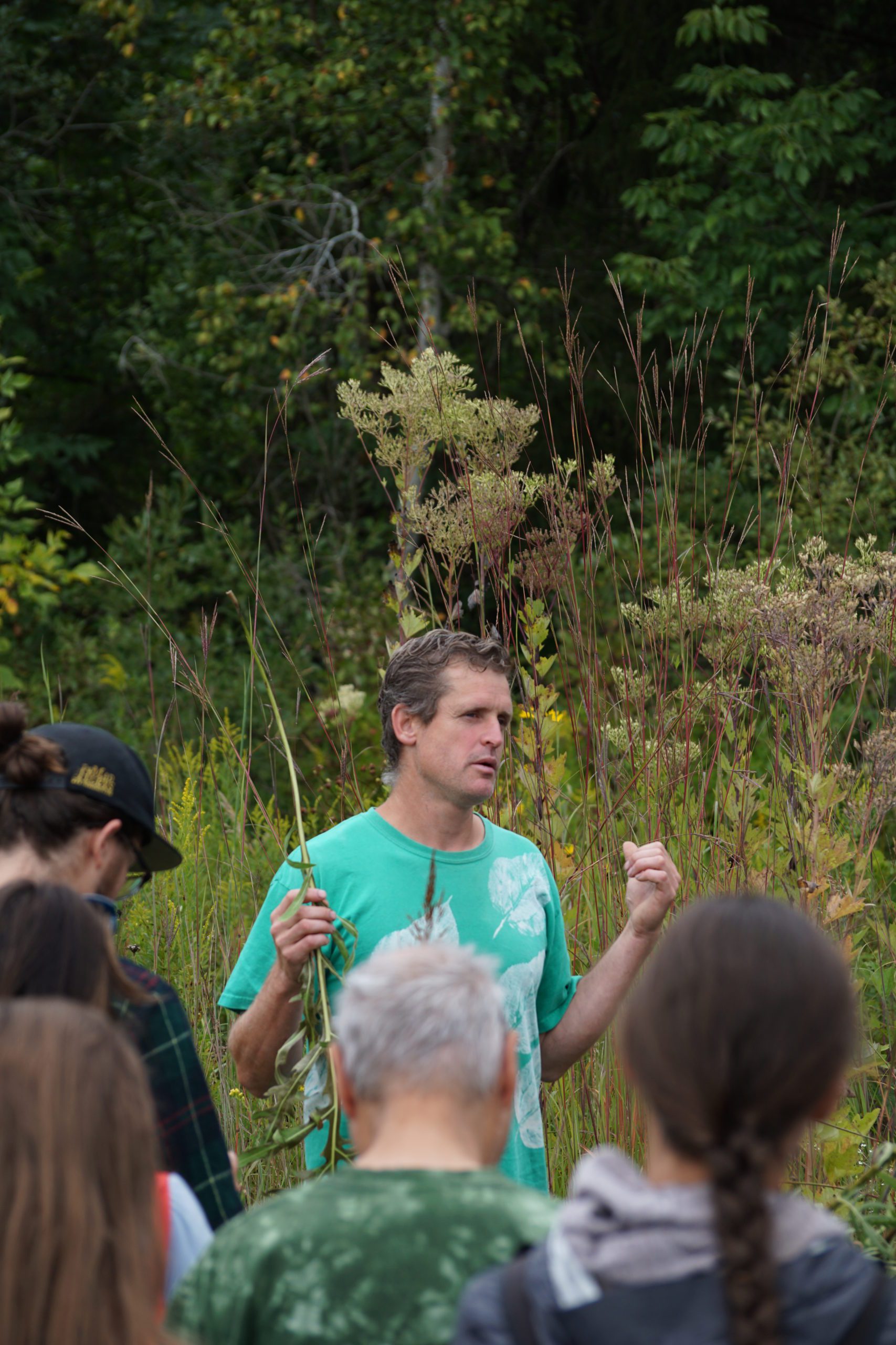 Sam Thayer, a white middle aged man, stands in a prairie holding a plant and speaking to a group of mixed aged adults