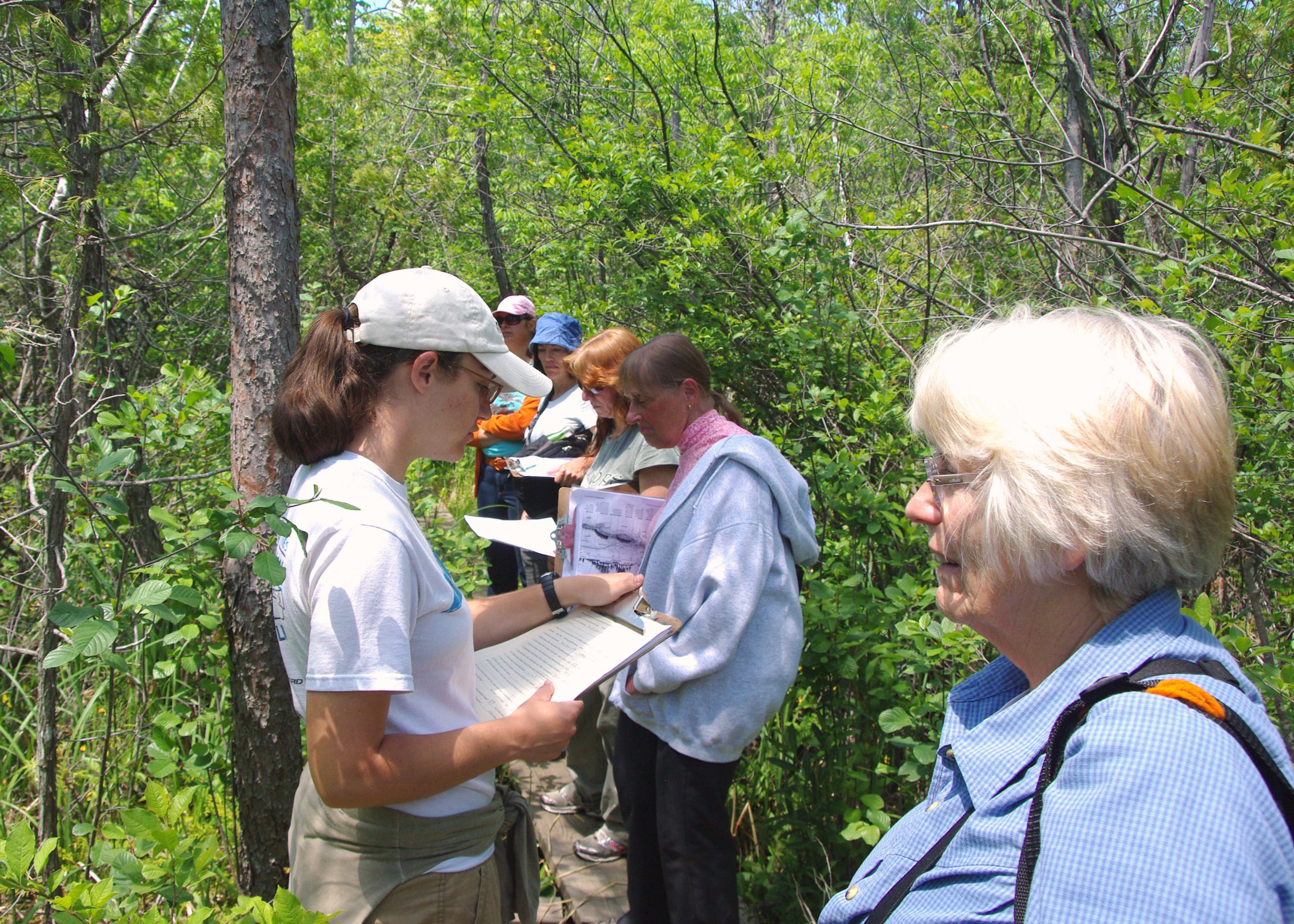6 people standing on a narrow boardwalk in the woods at the cedarburg bog looking at clipboards