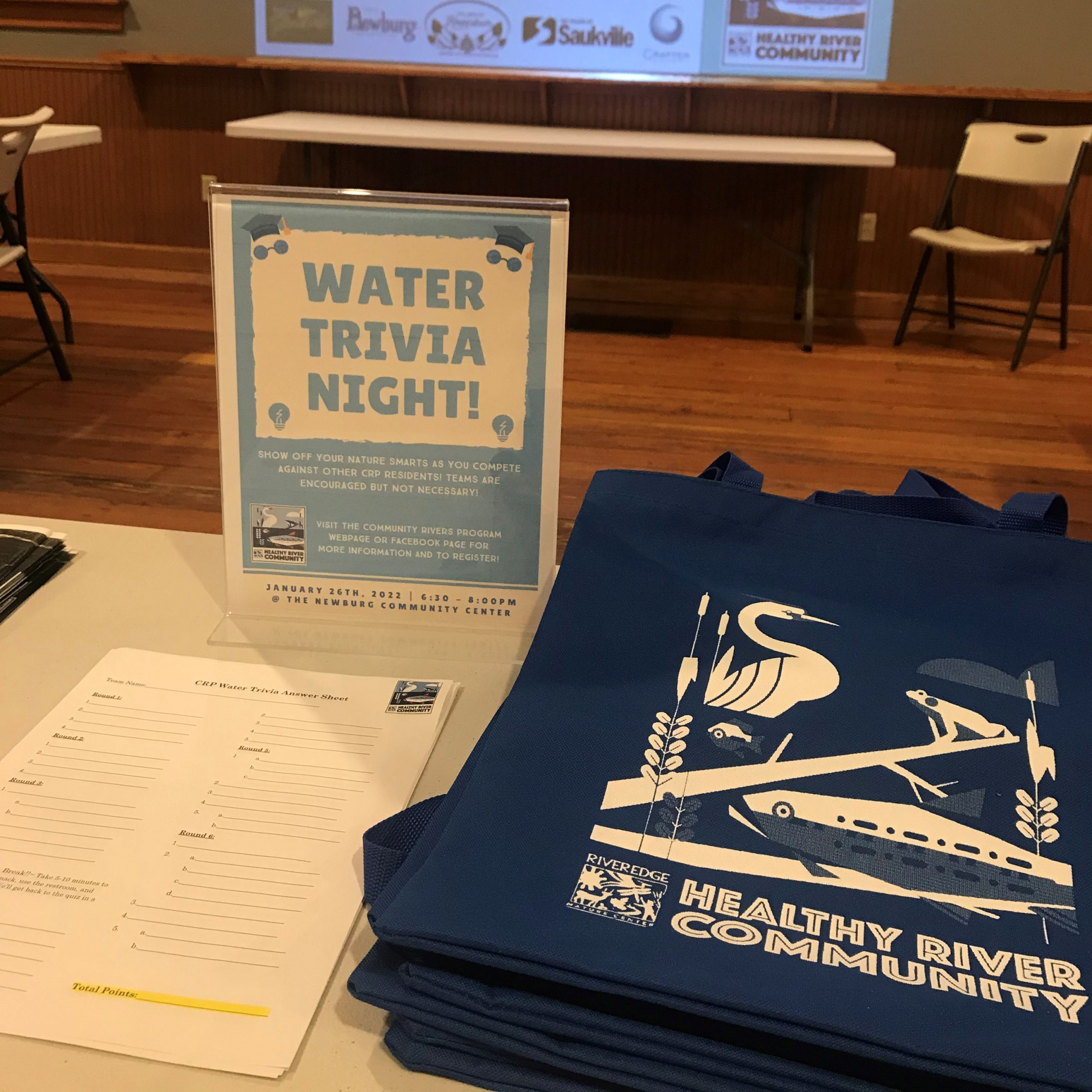 projector displaying "Trivia Night" with a light bulb icon. In the foreground is a table with another display for trivia night and a stack of dark blue "Healthy River Community" tee shirts