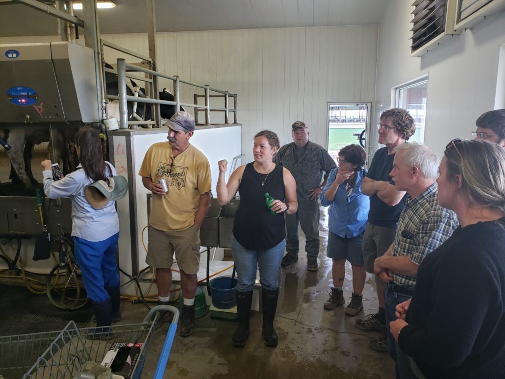 small group of people standing in a milking barn looking at a cow being milked