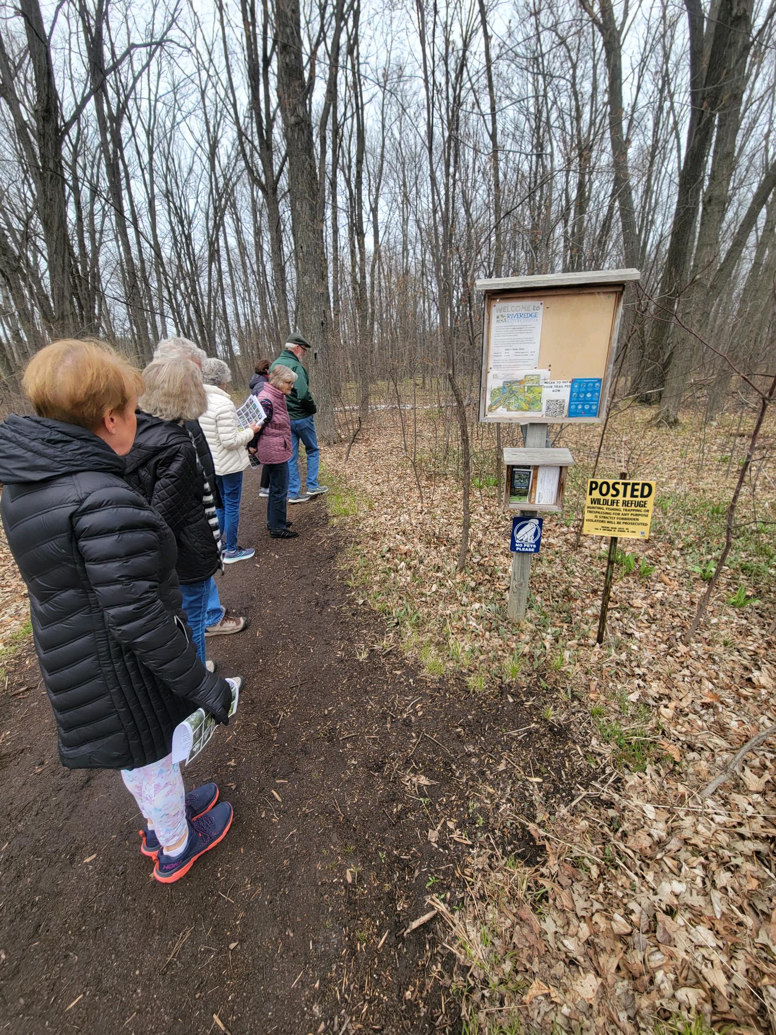 a group of older adults wearing coats stands on the dirt trail in the forest looking at a kiosk