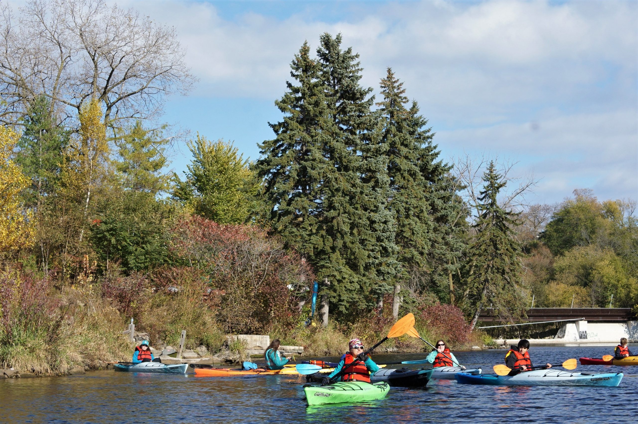 a small group of people paddling kayaks on the Milwaukee River in fall with evergreen trees in the background
