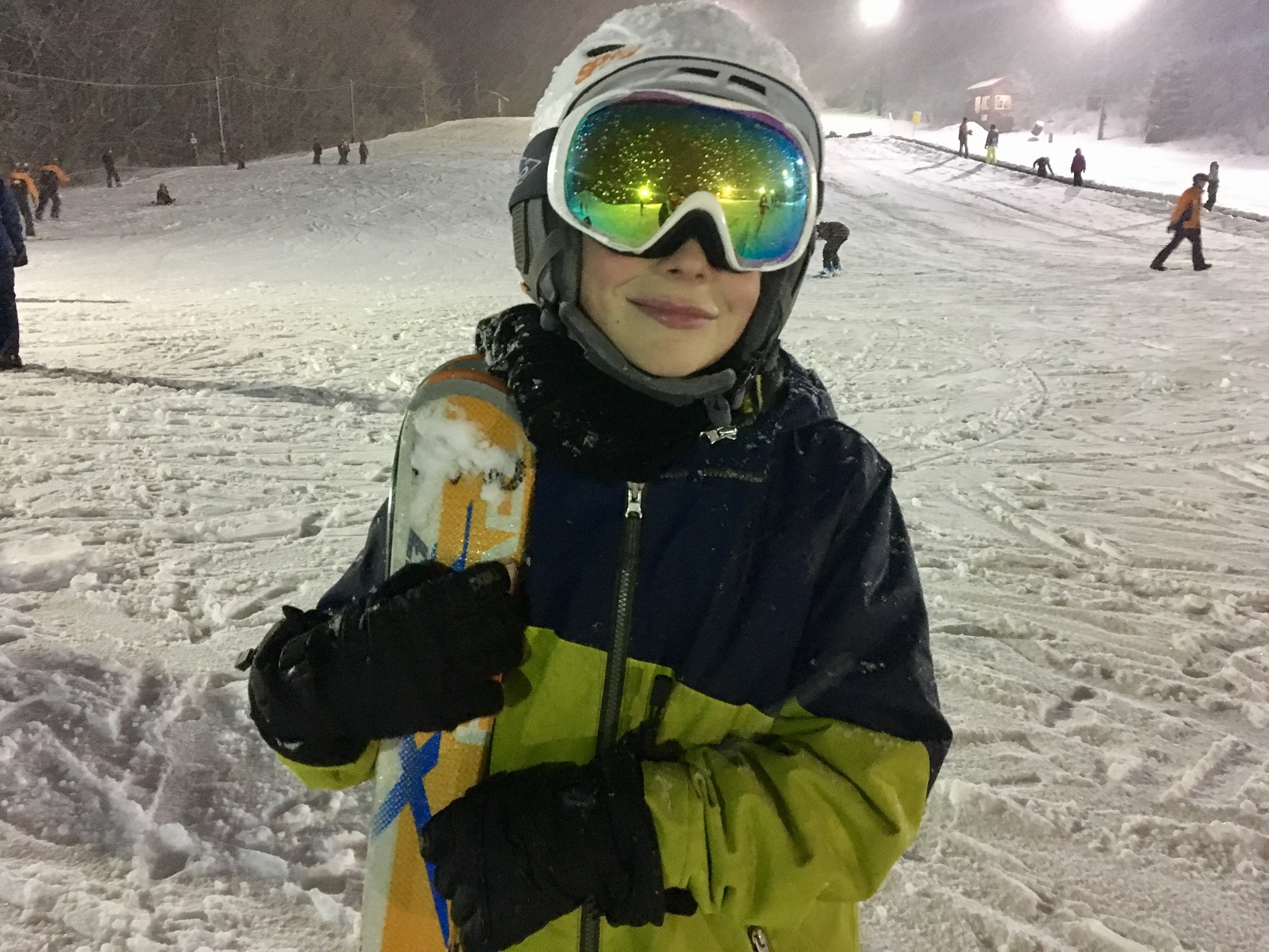 a child dressed in full snowboarding gear holds a snowboard and stands toward the camera with a ski hill covered in snow in the background