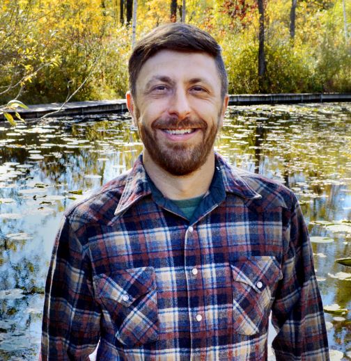John Rakowski, a middle aged white man with short brown hair, a mustache, and a short beard smiles at the camera with a sunny view of a pond in fall in the background