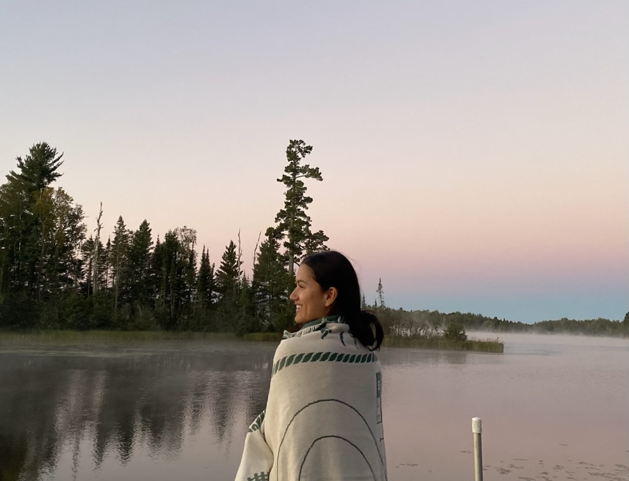 Erica Bhatti, citizen of the Lac du Flambeau Ojibwe, stands smiling looking at a mis covered lake with pines in the background