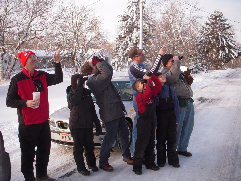 group of adults and kids dressed in winter gear stand on a street using binoculars to look for birds