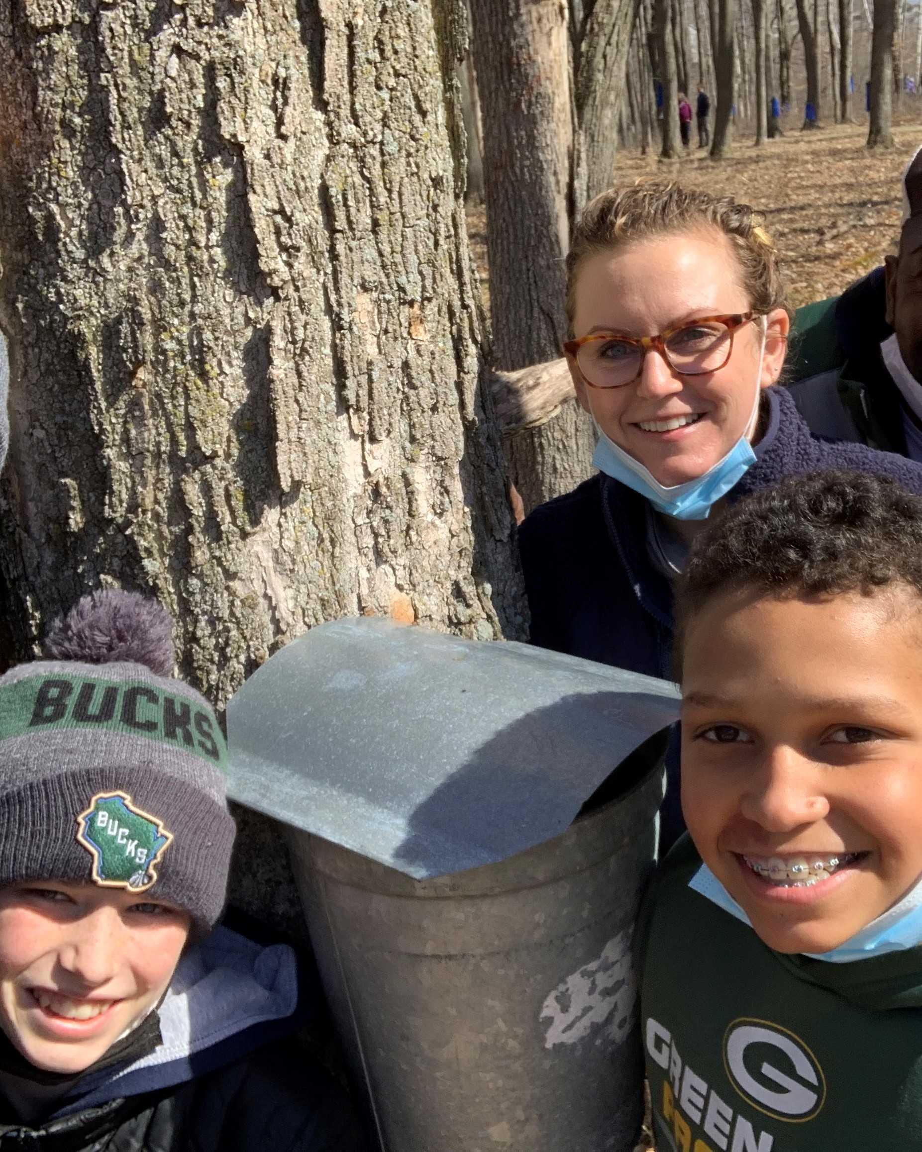 1 adult and 2 kids standing by a tapped maple tree and smiling at the camera