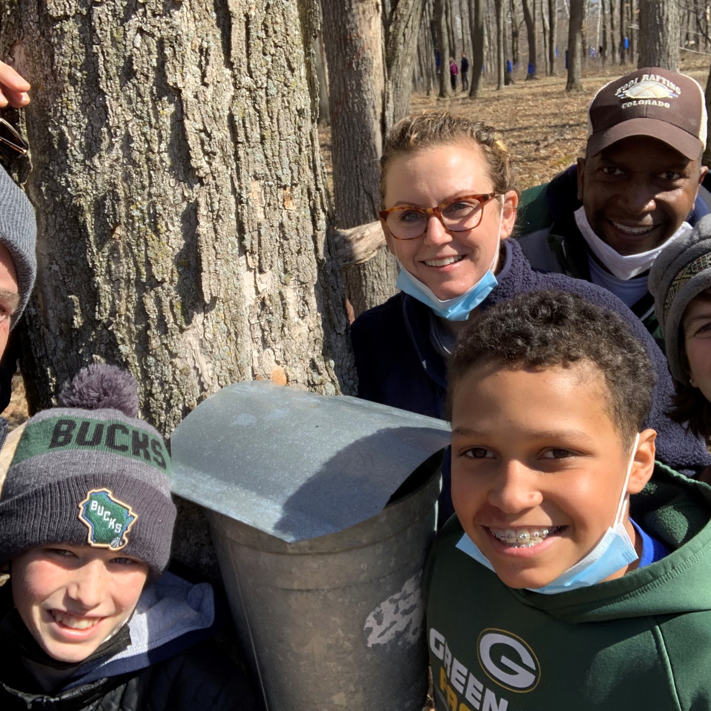 1 adult and 2 kids standing by a tapped maple tree and smiling at the camera