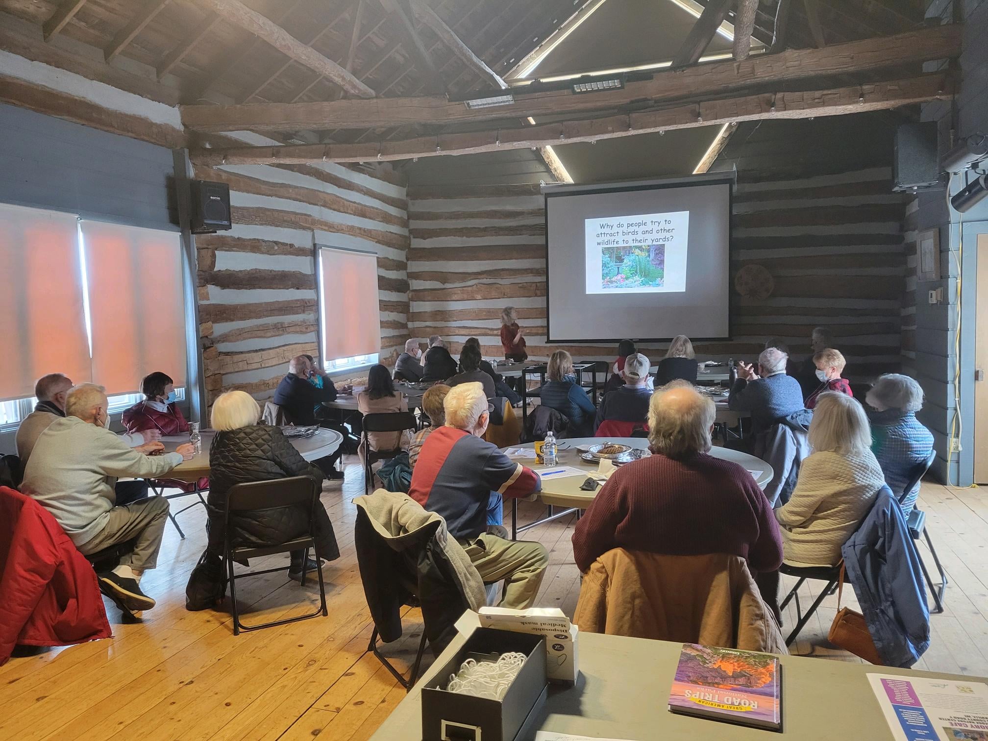 a group of senior adults sit at tables and chairs in the Riveredge Barn watching a presentation on a projector