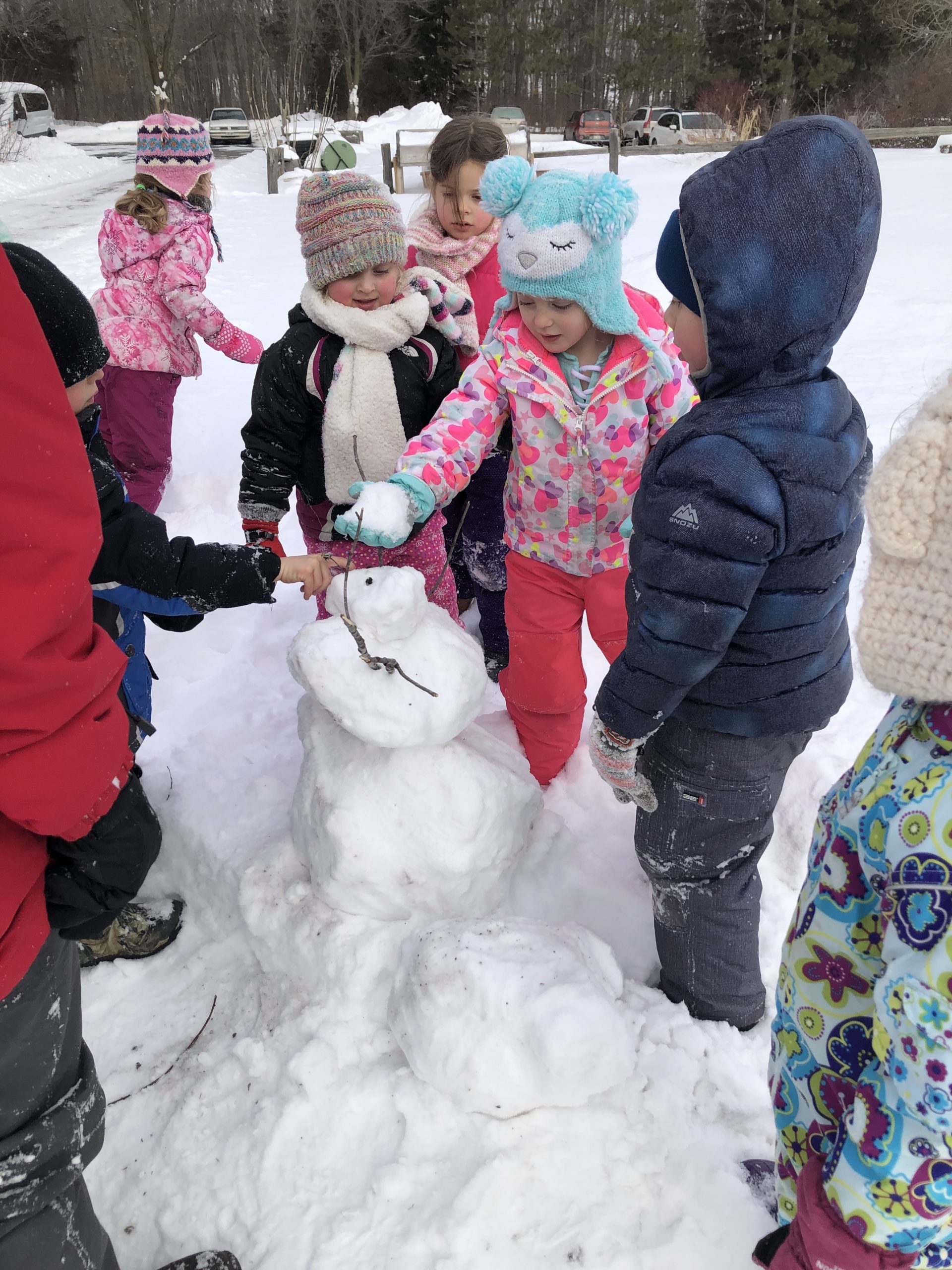 a small group of young children dressed in winter gear build a snowman in the snow