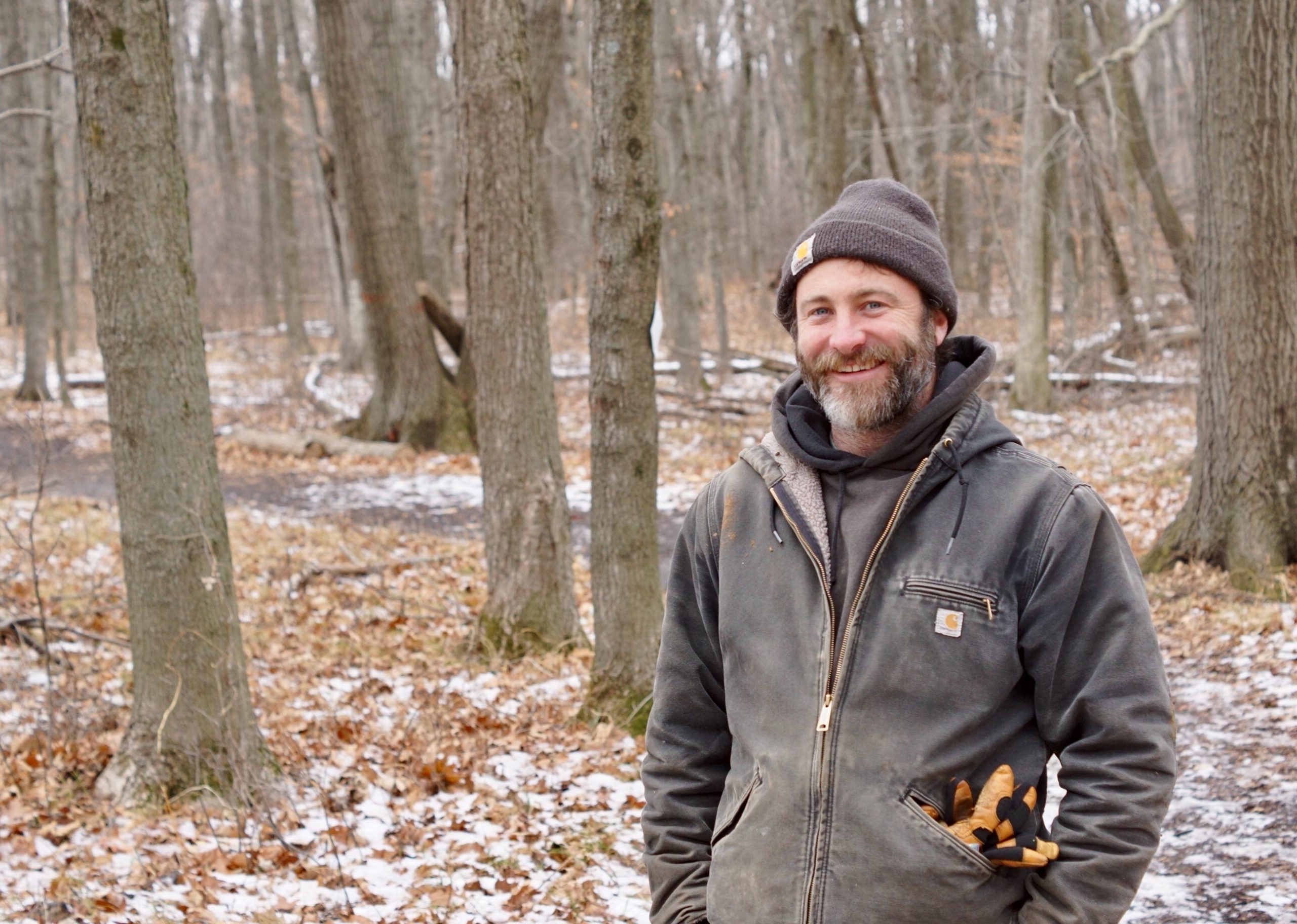 Joe Mantoan Jr, a middle age white man with facial hair, stands in the Riveredge forest with a dusting of snow on the ground while smiling at the camera