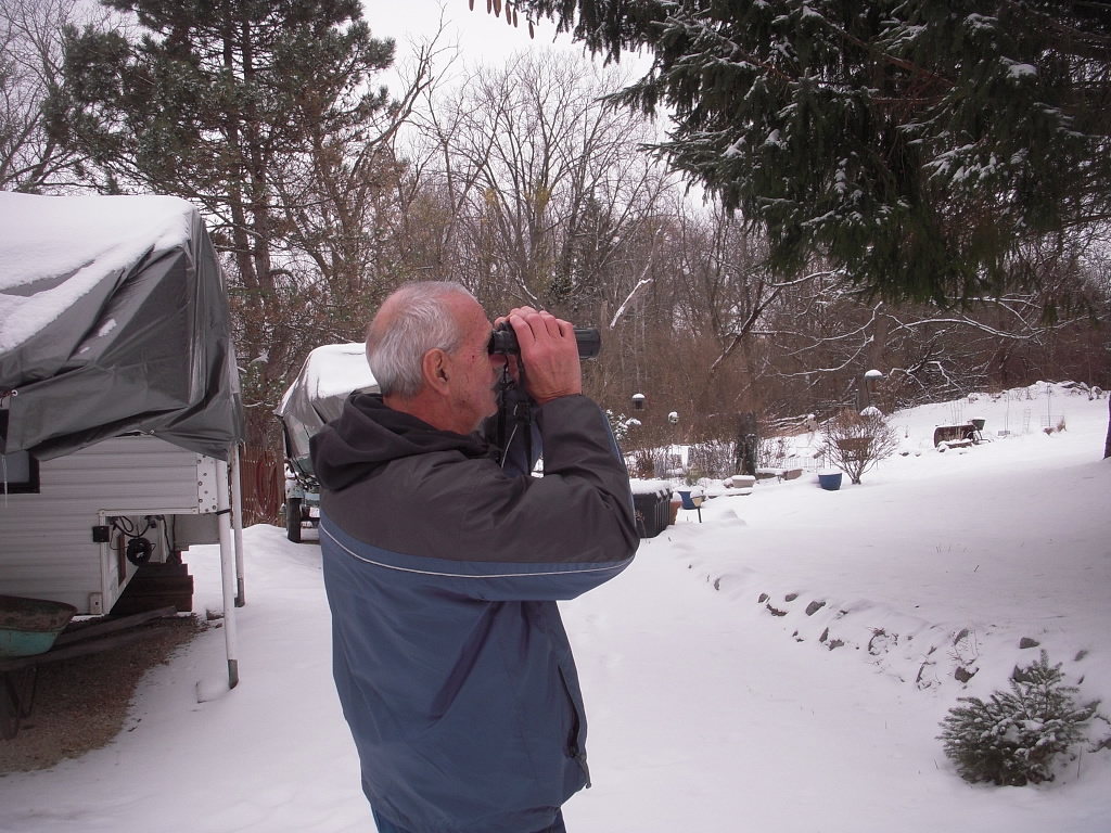 Steve Kupcho, an older white man with short white hair dressed in winter gear, stands outside on a winter day using binoculars to look off into the distance