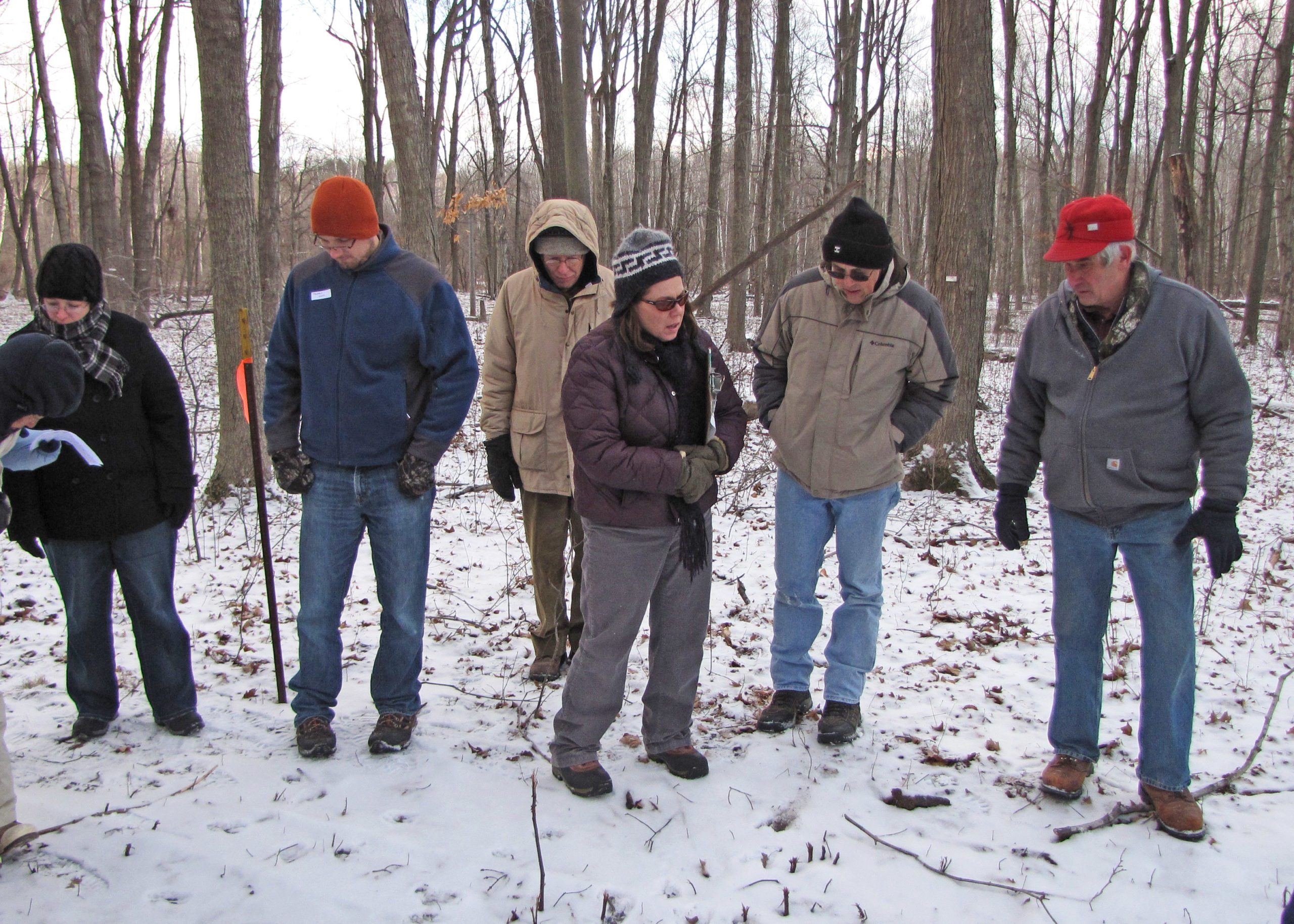 6 adults dressed in winter gear stand in the Riveredge woods with snow on the ground looking at a speaker