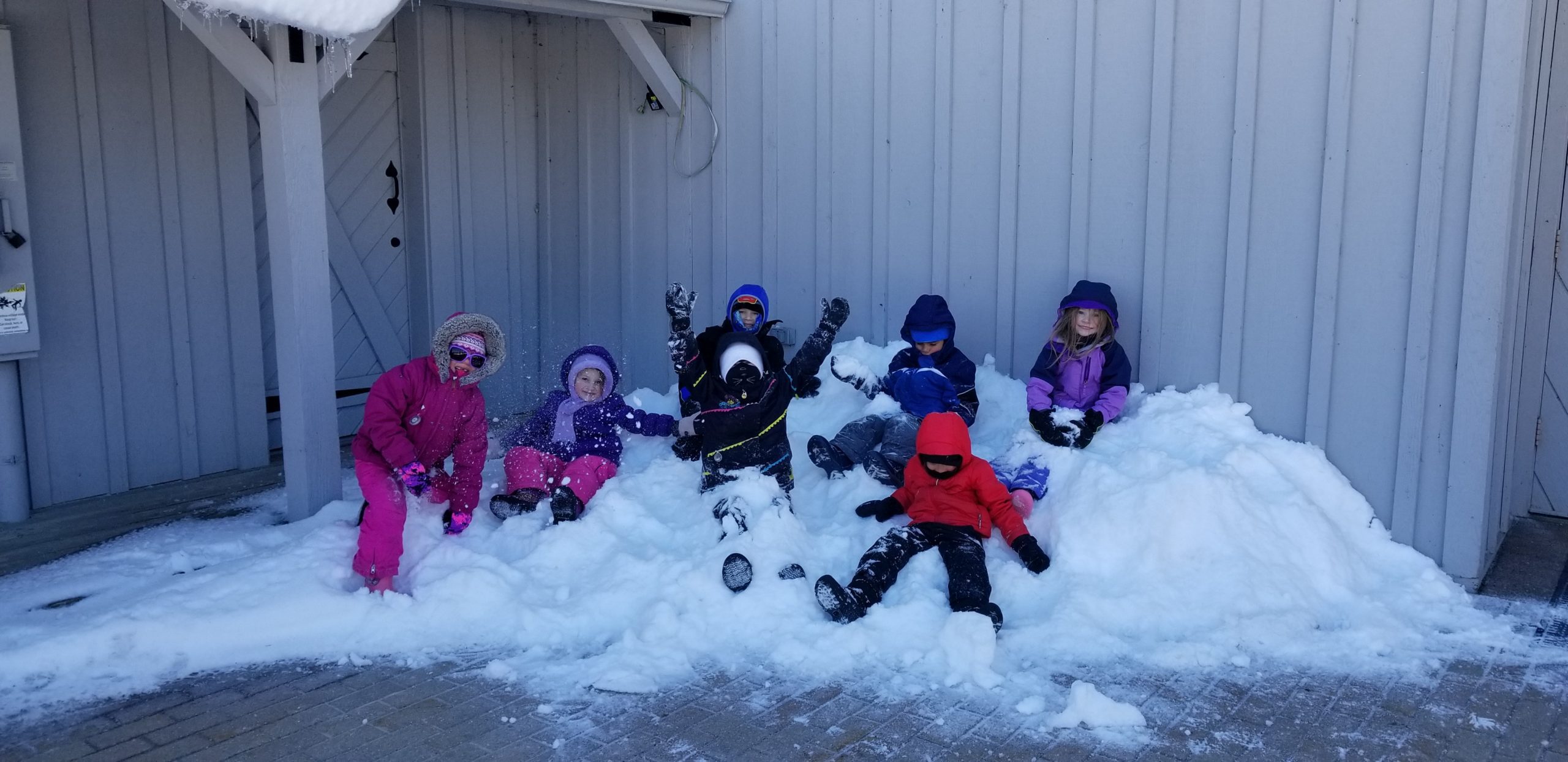 7 young kids dressed in winter gear sit on a pile of snow