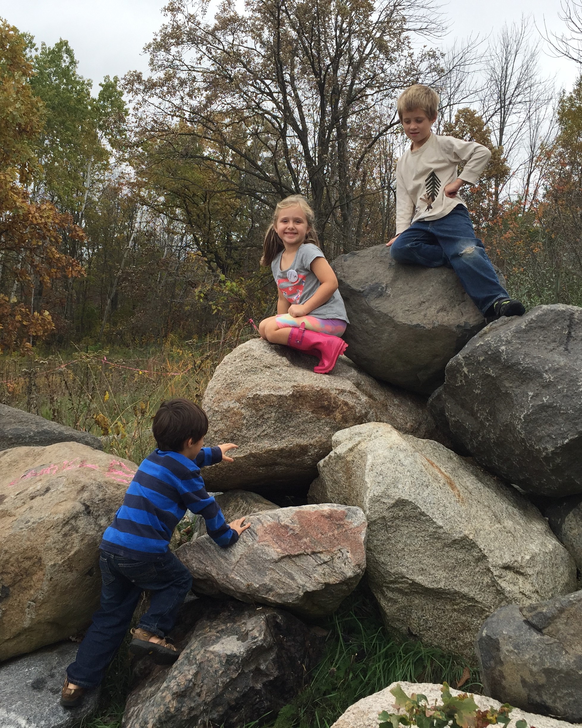 3 young kids are climbing on the Larsen Climbing Rocks at Riveredge. One of them is smiling at the camera