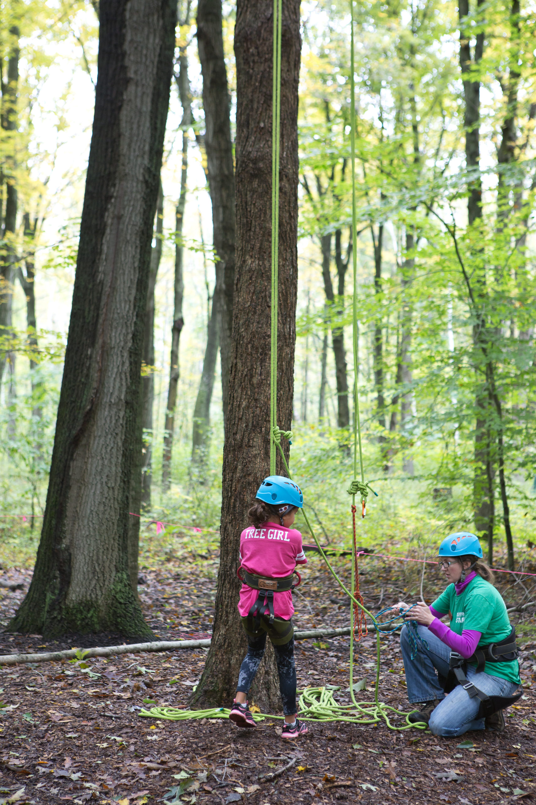 a child stand on the ground in a harness attached to a tree climbing rope listending to an instructor before climbing