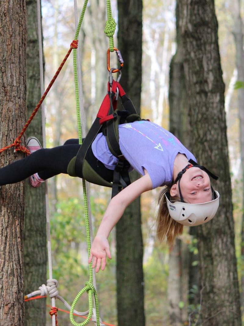 a young child smiels at the camera while hanging upside down from a tree in a harness with tree climbing ropes