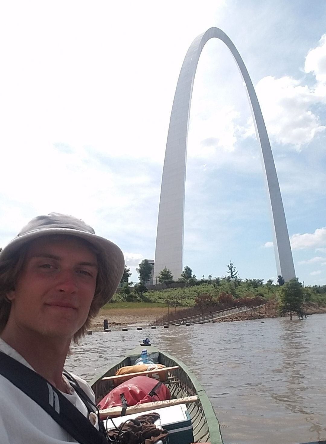 Luke Yourzak, a young white man, smiles at the camera while sitting in a canoe on the Mississippi River with the St Louis arch in the background