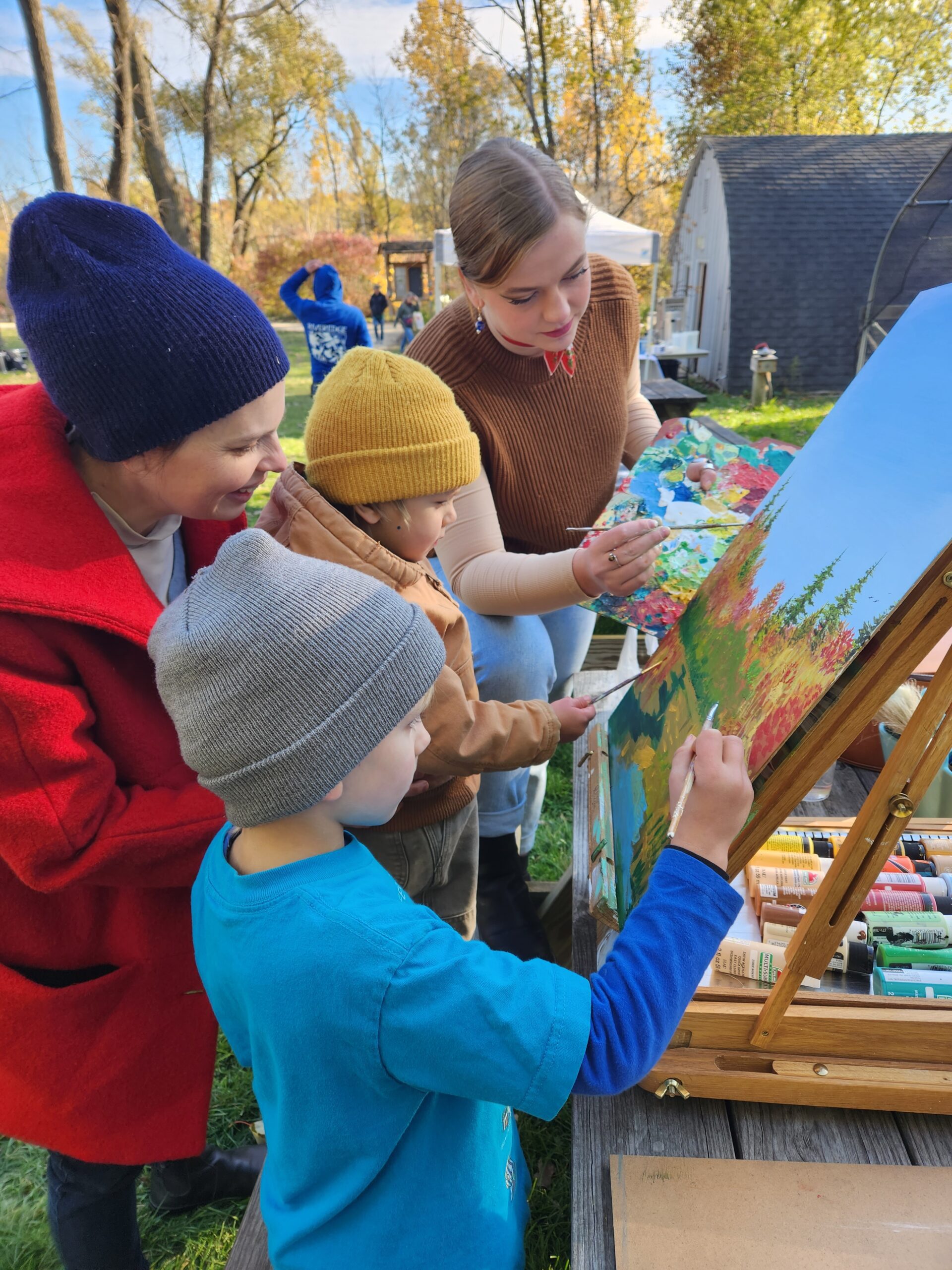 2 young kids in fall attire assited by an artist and an adult paint fall trees on a large cavas