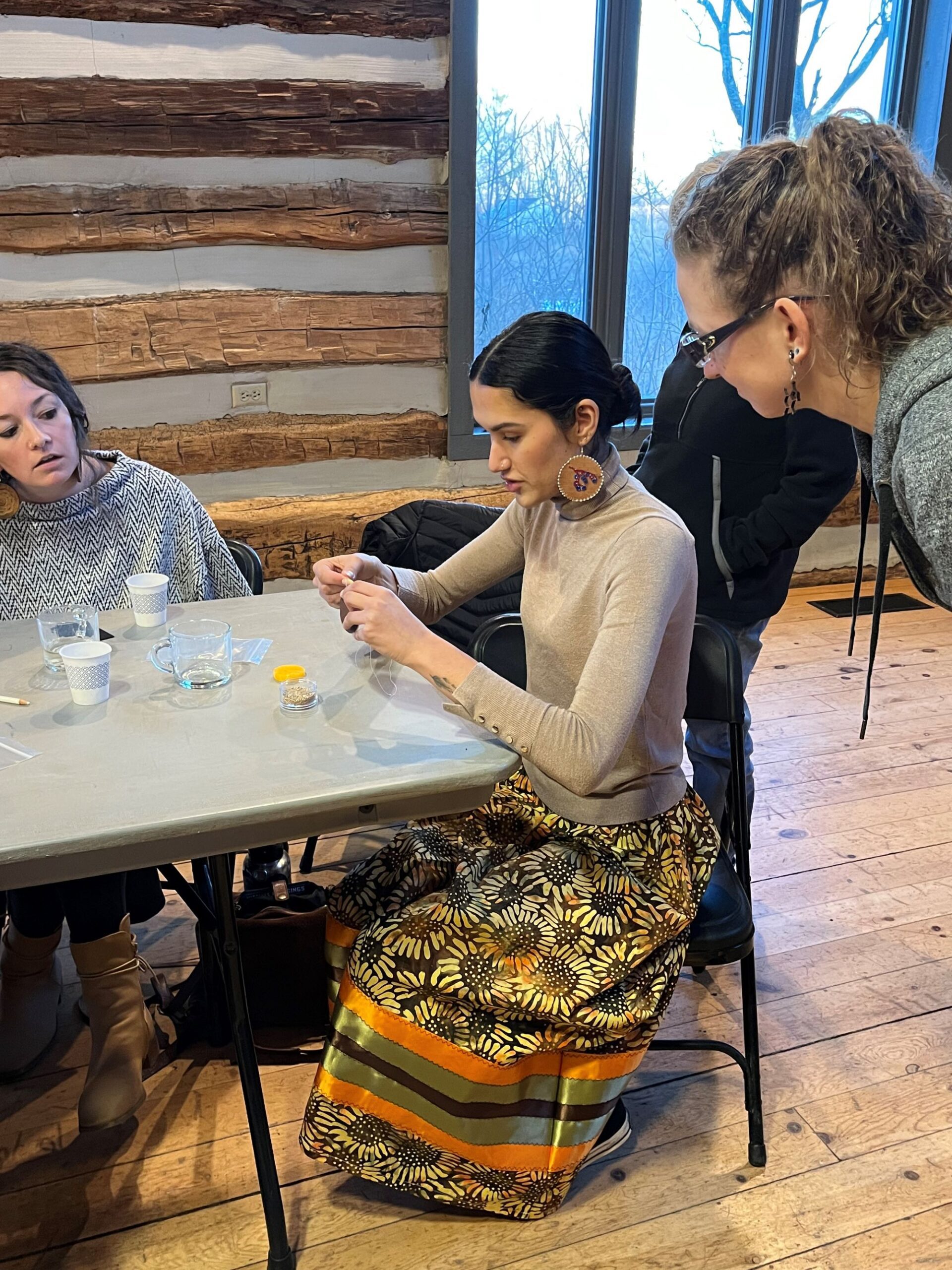Erica Bhatti sits at a table demonstrating beadwork techniques to 2 adults looking on
