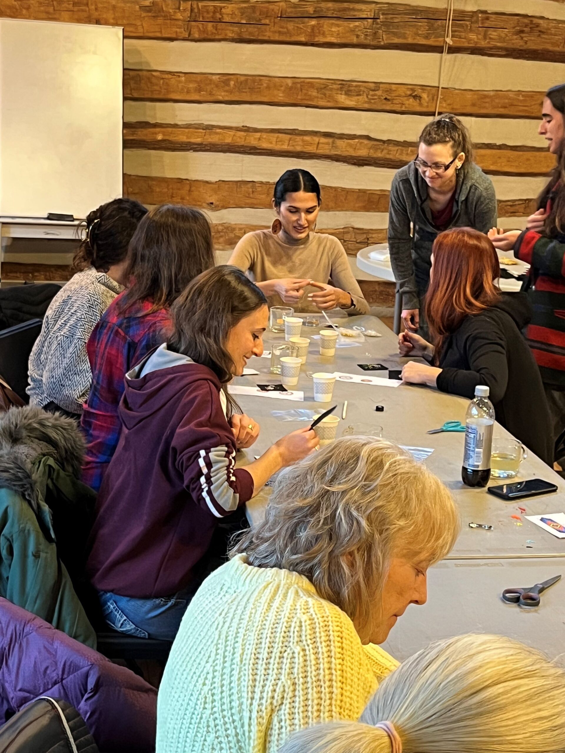 Erica Bhatti sits at a table demonstrating beadwork techniques to a small group of adults looking on and practicing