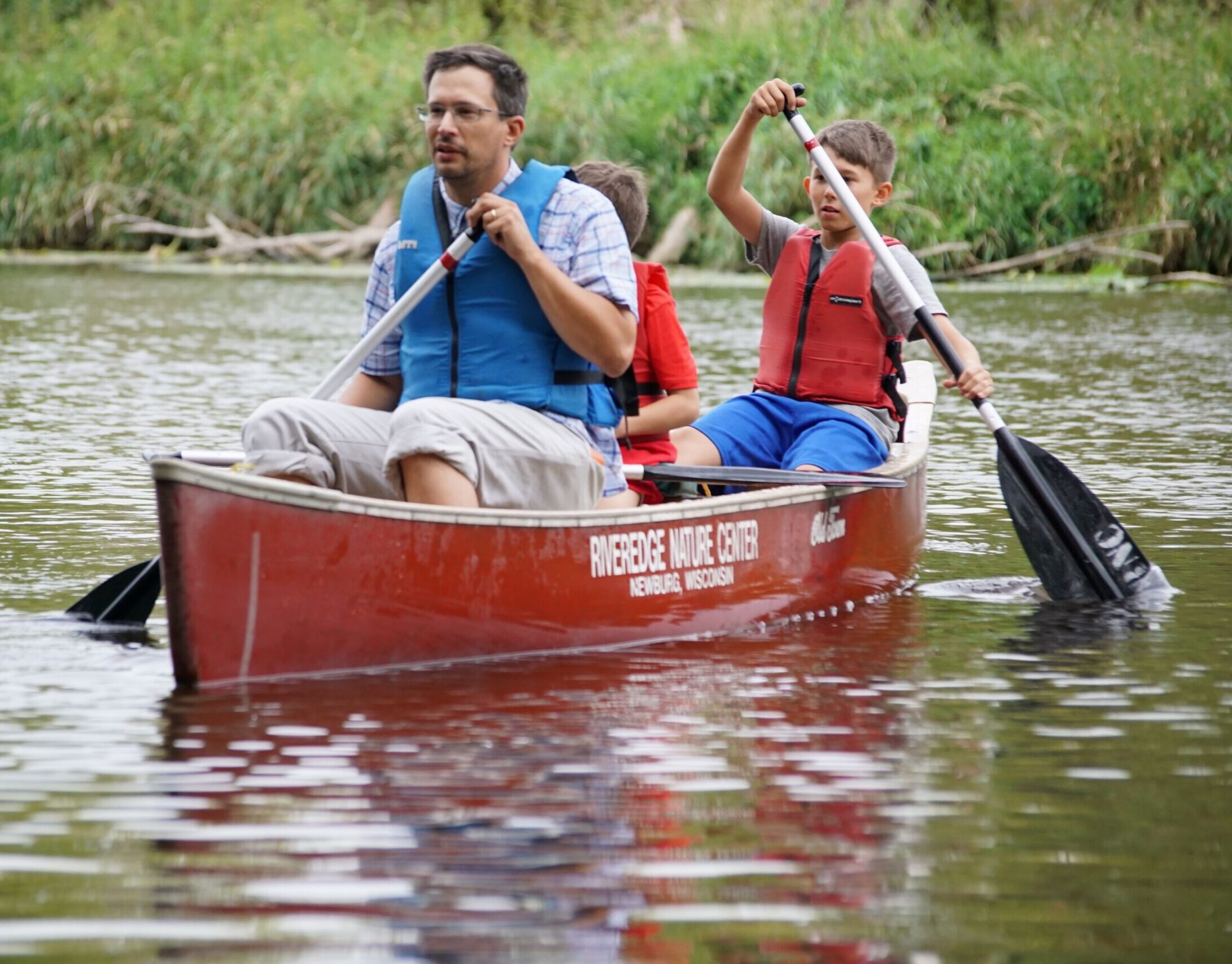 a parent and two children paddle in a red canoe with cattails in the background