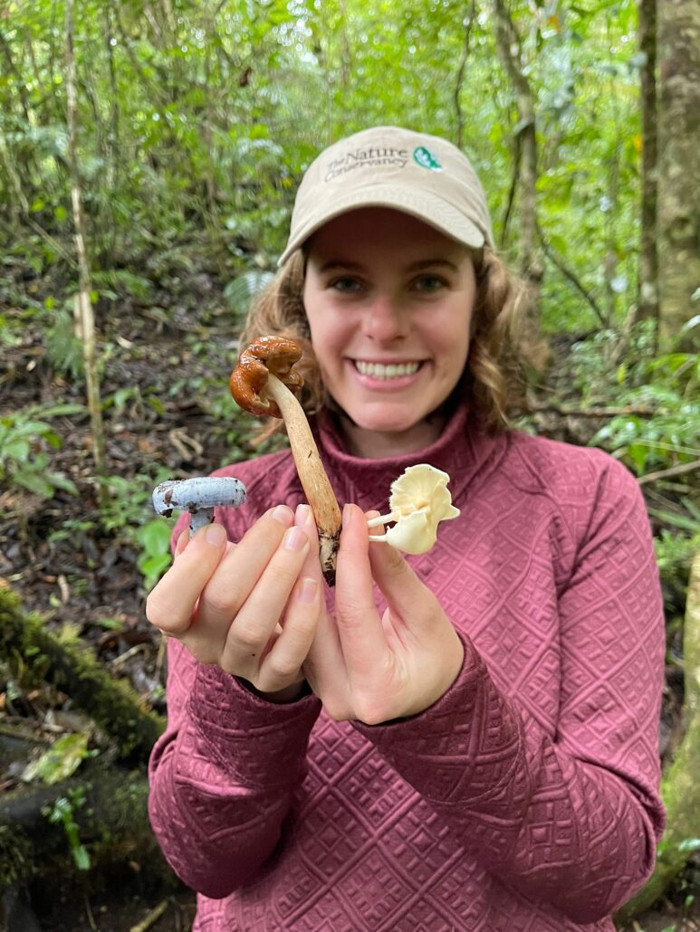 Oliva, a young white woman, smiles at the camera while holding 3 different types of mushrooms