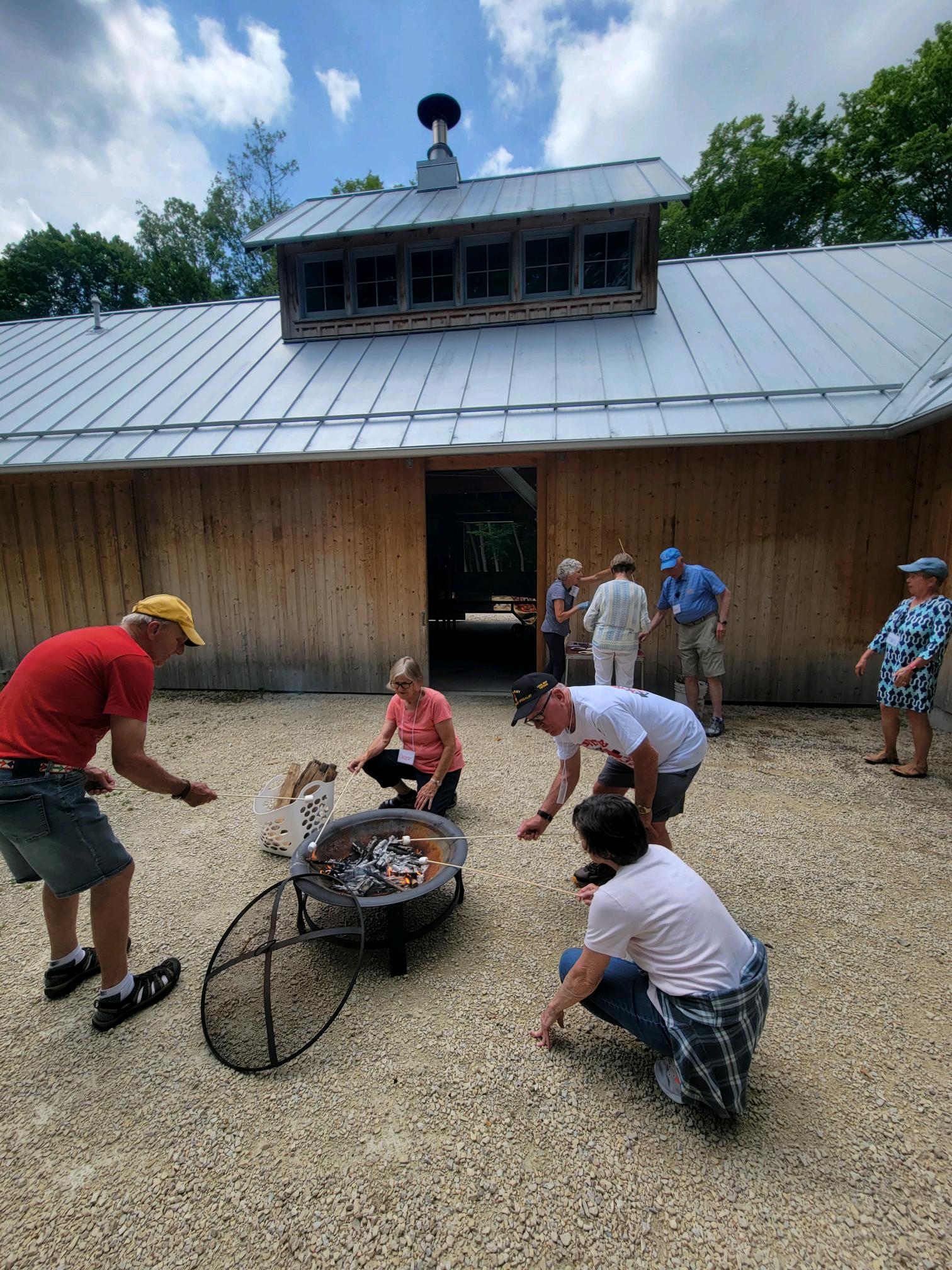 4 older adults kneel around a campfire by the Riverdge Sugarbush House roasting marshmallows on sticks