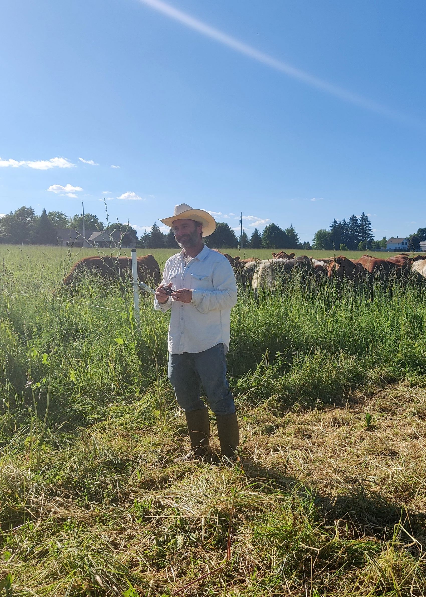 Joe Mantoan, a middle age white man wearing a cowboy hat, stands and smiles with his brown cows in the background on a sunny day