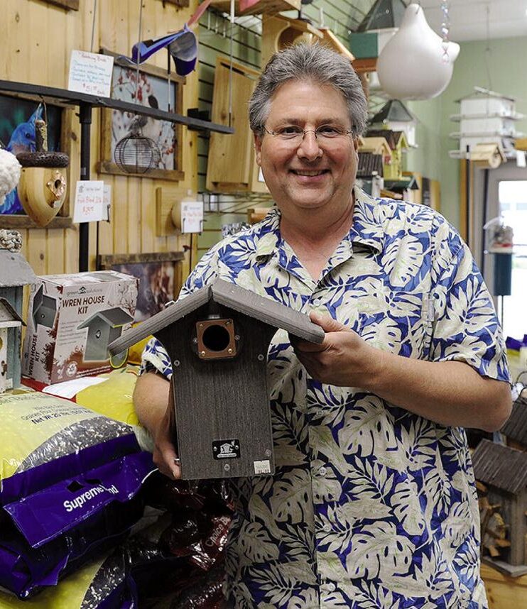 Dan Panetti, a white man with gray hair, smiles at the camera while holding a wooden bird house with other bird supplies behind him