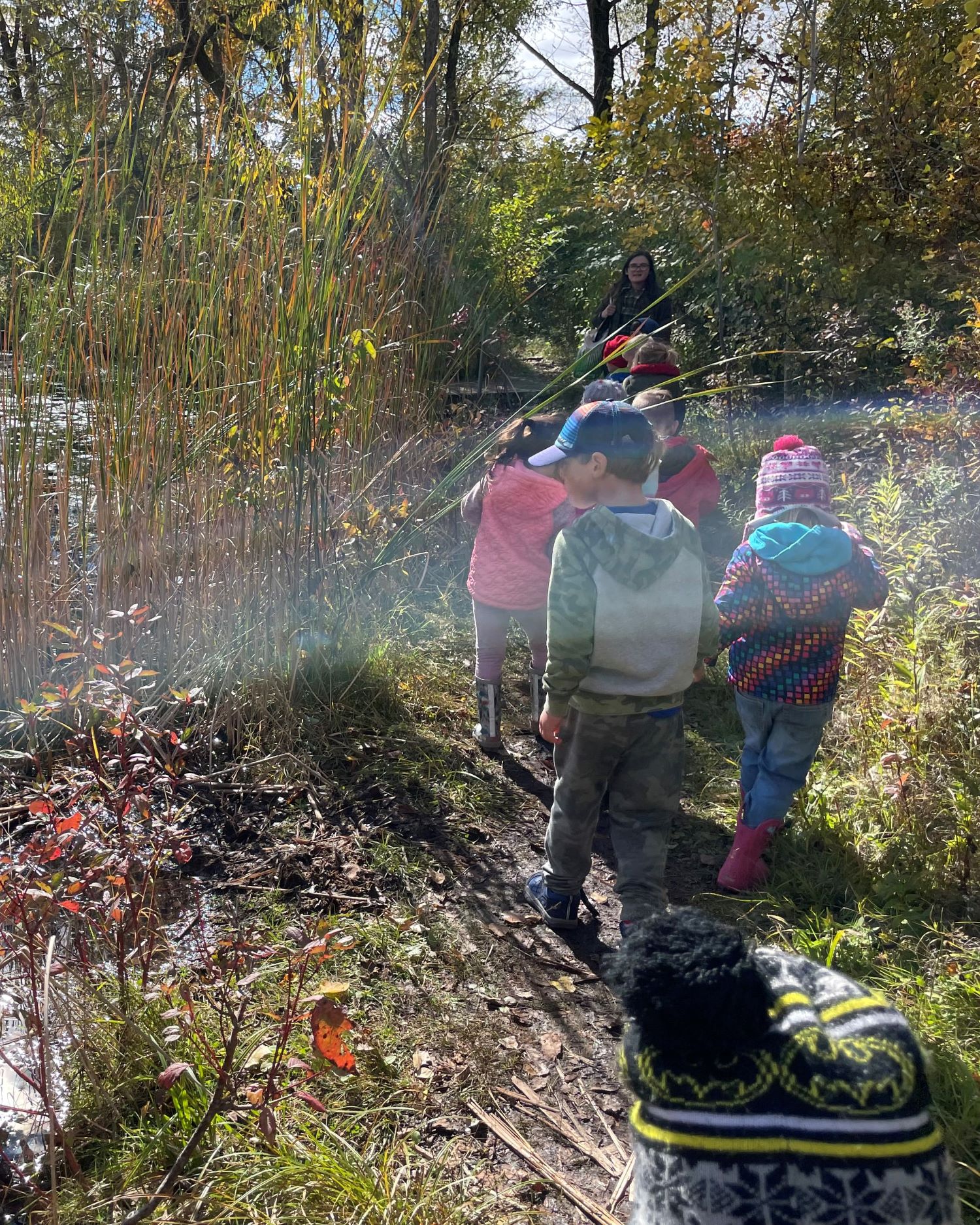 a small group of young kids walks on a trail next to a pond in fall