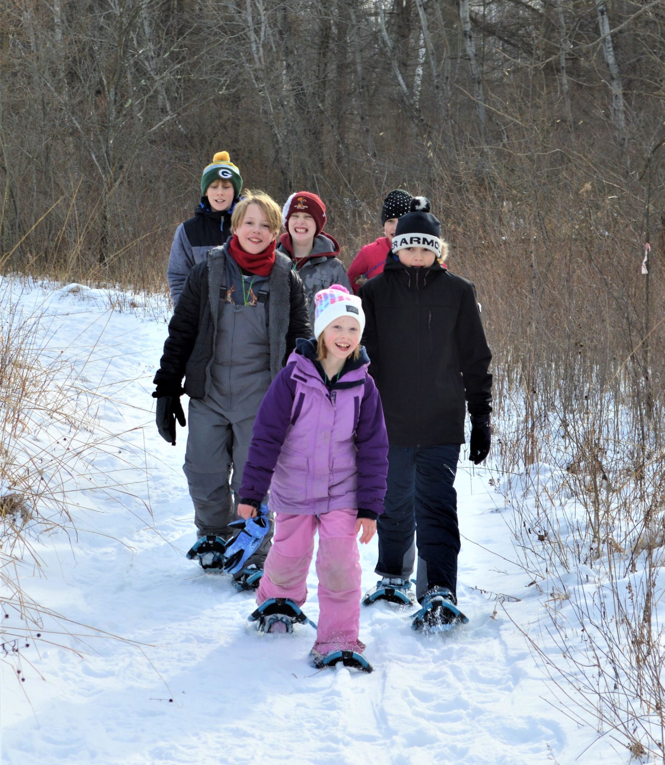 6 kids of varying ages dressed in winter gear snowshoeing on a snow covered trail through the Riveredge prairie