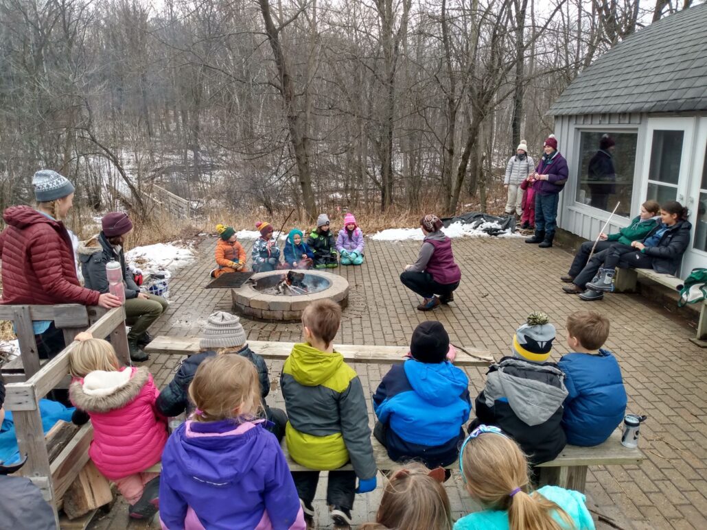 large group of winter campers gathered around a small campfire listen to a speaker