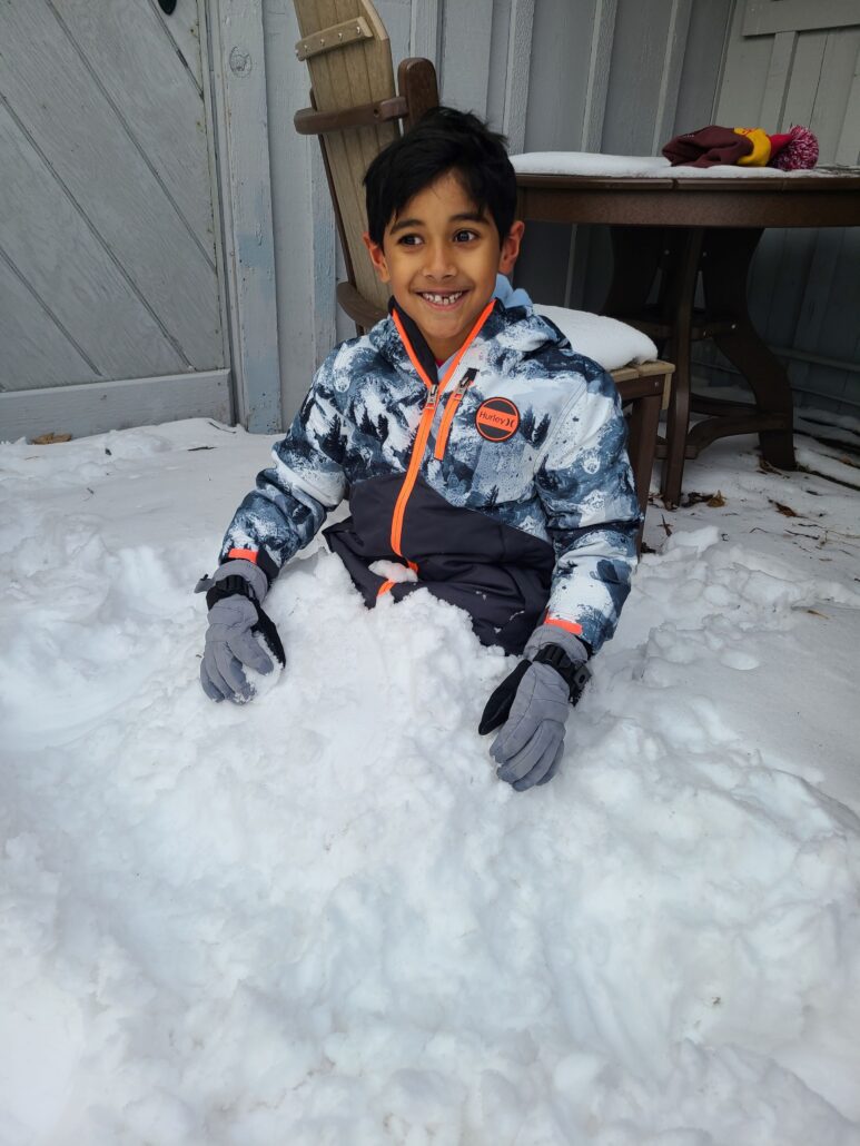 a young child smiles at the camera. He has buried himself in a pile of snow outside the Riveredge barn.