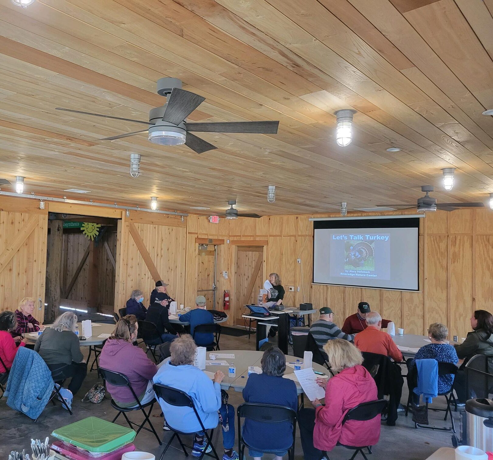 a group of older adults sits in the Riveredge Sugarbush House watching a presentation on a screen about Turkey given by Mary, a Riveredge educator