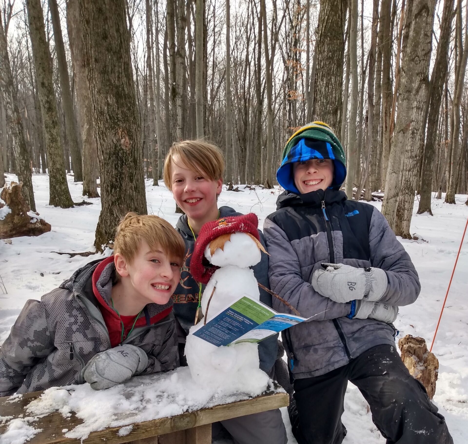 3 kids bundled up in the Riveredge forest in winter smile at the camera with the snowman on a bench in the center