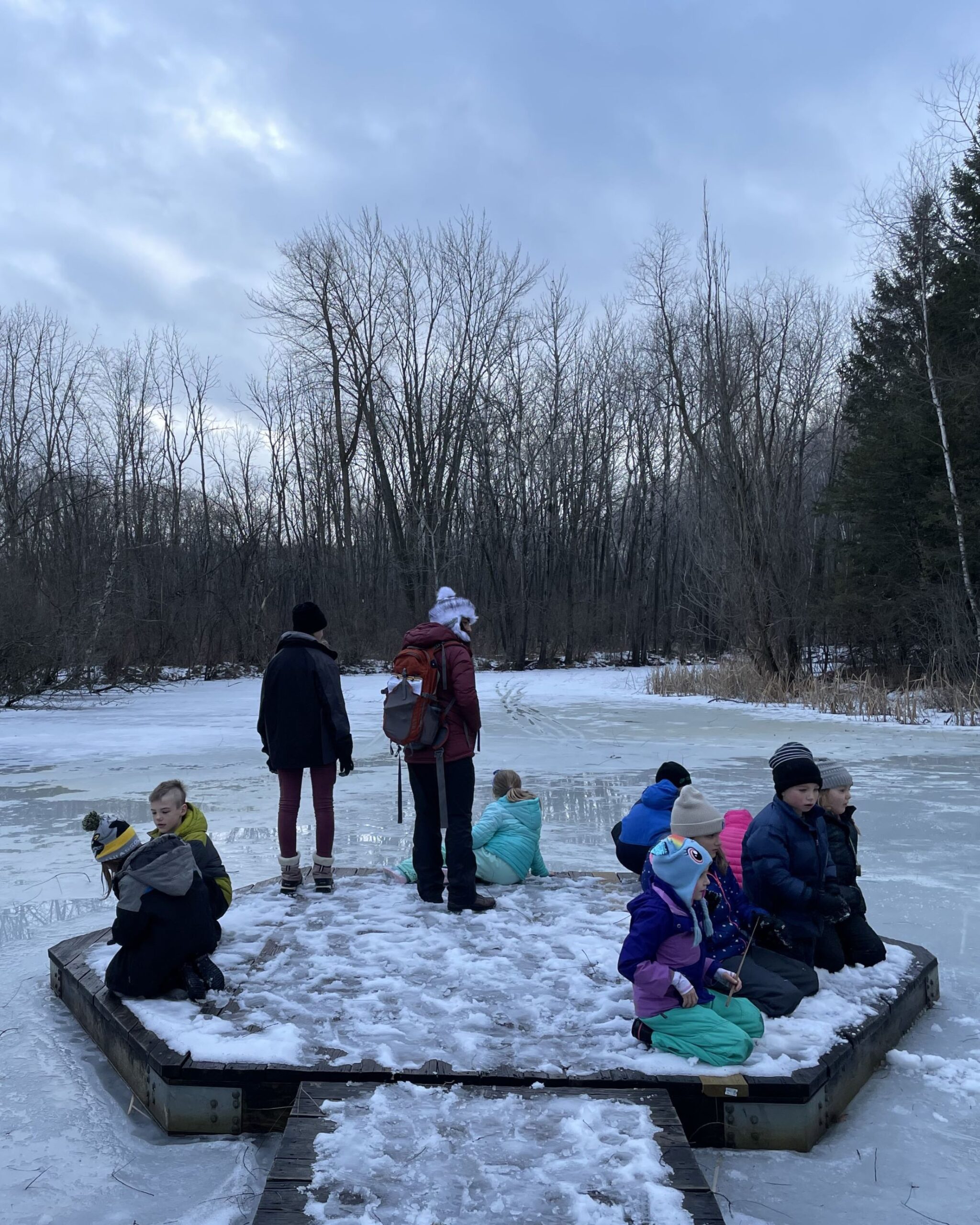 a small group of kids and adults are on a wooden pond dock at Riveredge looking at the frozen pond around them