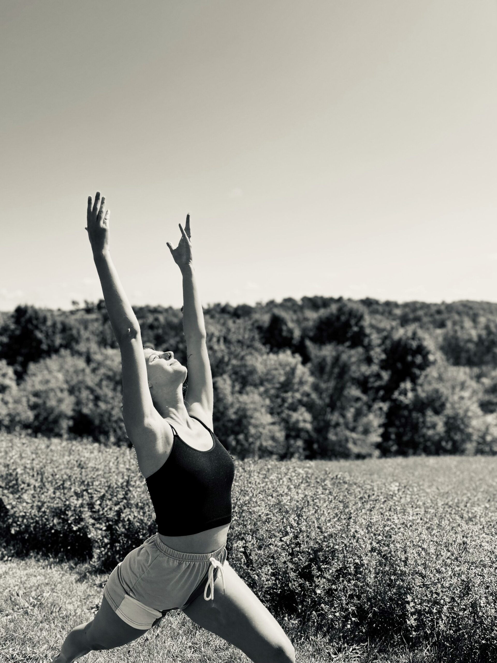 black and white photo of Mary Addy, a young white yoga instructor, doing a yoga pose reaching toward the sky with grass and trees in the background