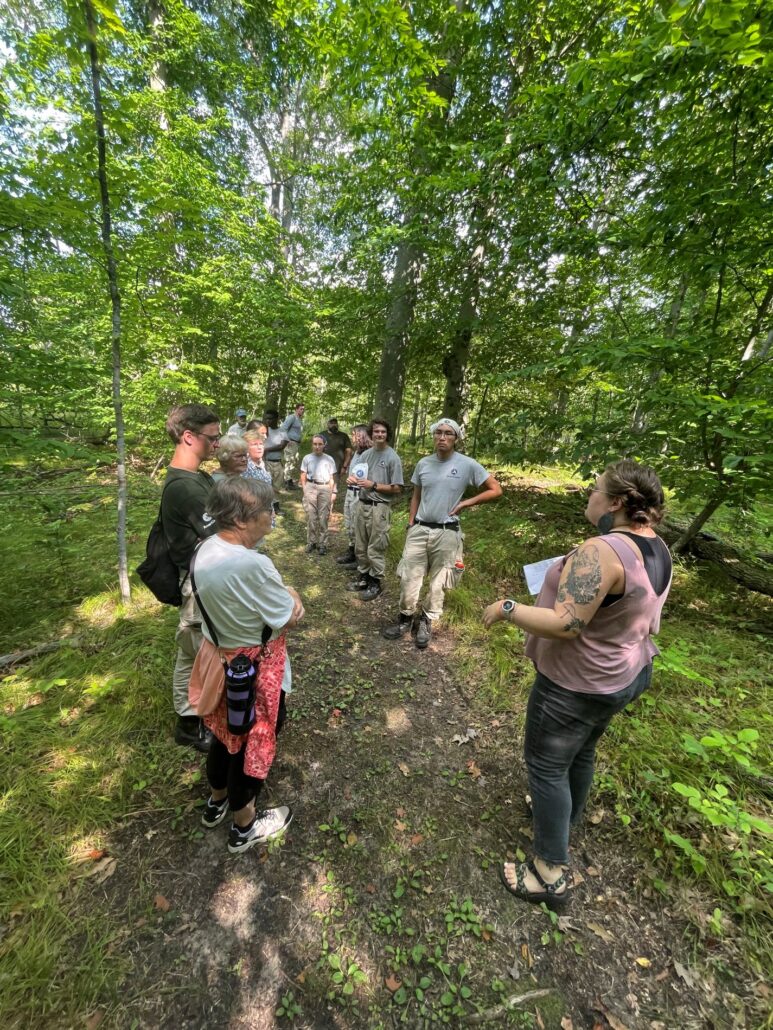 a small group of adults standing on a trail in the Riveredge forest listening to a speaker
