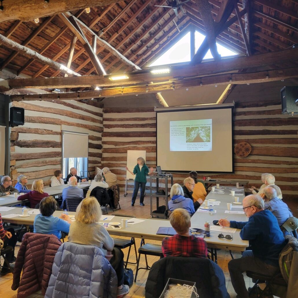 a group of older adults sitting at table in the Riveredge barn watching a slideshow about Christmas trees