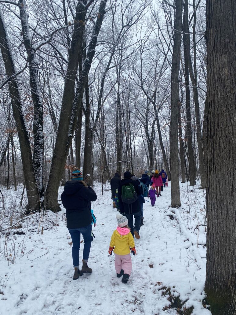 a small group of young children and adults take a walk through the Riveredge woods in winter with snow on the ground